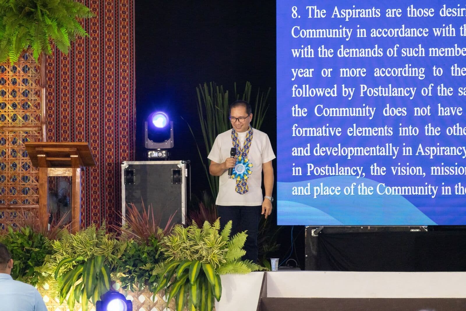 A speaker on a stage beside a screen showing a passage about the Community’s Aspirants