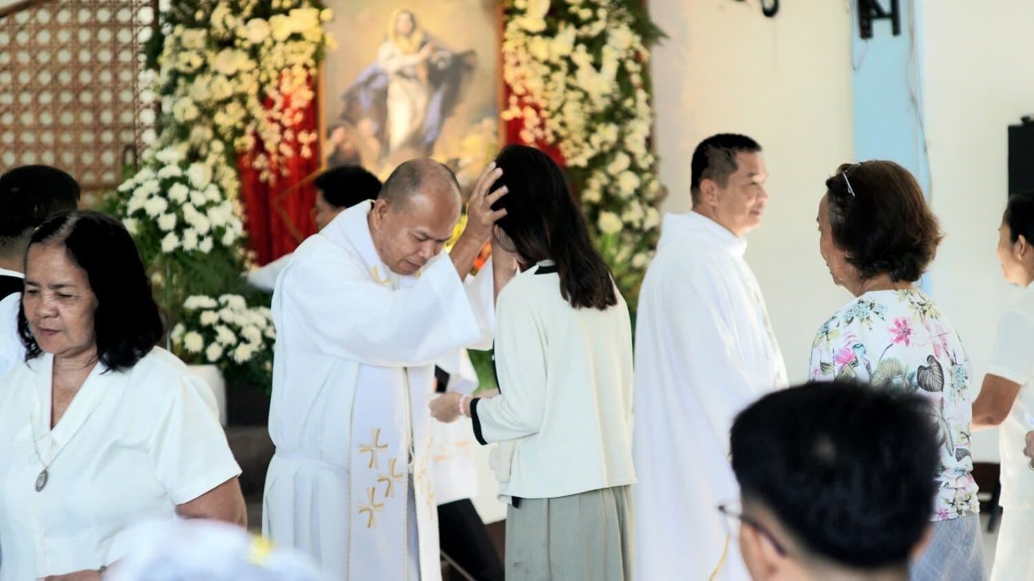 A priest in white vestments laying his hand on a woman’s head in blessing, others waiting beside him