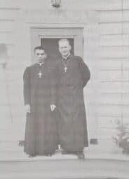 The young Fred Kriekenbeek in a black cassock, standing beside another priest at a church door