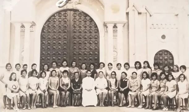 An old sepia photograph of the young Fr. Fred in a white cassock, seated among about forty women in front of the great wooden doors of a church.